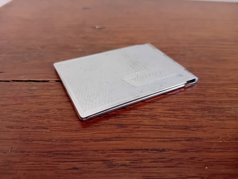 May include: A silver metal compact with a textured surface and the word "Germany" engraved on the front.