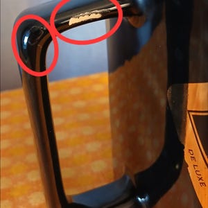 May include: Close-up of a black mug with a glossy finish. The handle shows signs of wear with chipped paint, revealing the underlying material. A partial view of a label with the word "DELUXE" is visible.