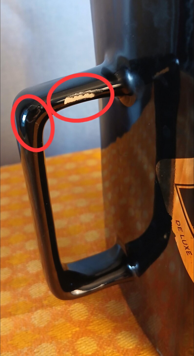 May include: Close-up of a black mug with a glossy finish. The handle shows signs of wear with chipped paint, revealing the underlying material. A partial view of a label with the word "DELUXE" is visible.