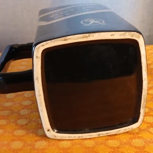May include: Black ceramic mug with a square base and a handle. The mug has a white rim and a dark blue section with text and a logo. The mug is sitting on a yellow and orange patterned surface.