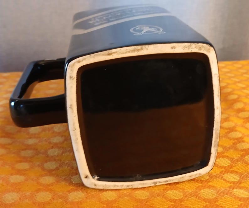 May include: Black ceramic mug with a square base and a handle. The mug has a white rim and a dark blue section with text and a logo. The mug is sitting on a yellow and orange patterned surface.