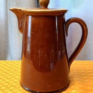May include: A brown ceramic pitcher with a lid and handle. The pitcher has a spout for pouring and a rounded knob on top of the lid. The ceramic has a glossy finish and a dark brown color.