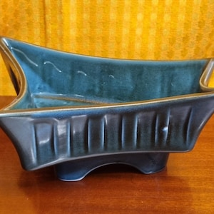 May include: A teal and navy blue ceramic planter with a unique, angular design. The planter has a boat-like shape with a textured exterior and a smooth interior. It sits on three small feet and is suitable for small plants or decorative use.