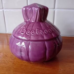 May include: A purple ceramic beetroot-shaped container with a lid. The word "Beetroot" is written on the lid. The container is sitting on a wooden surface.