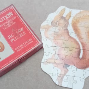 May include: A white jigsaw puzzle of a brown squirrel with a bushy tail. The puzzle is in a red box with the text "Squirrel Nutkin Beatrix Potter Famous Character Jig Saw Puzzle Cut-out Figure Size 14" x 11" inches".