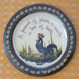 May include: A circular ceramic plate with a dark green border and a cream-coloured centre. The plate features a painted rooster and the text "A pound of pluck is worth a ton of luck."