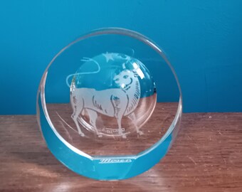 May include: A clear glass paperweight with a white etched image of a bull, the astrological sign for Taurus. The paperweight has a blue base and the word "Taurus" is etched on the bottom.