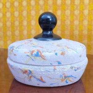 May include: A vintage powder box with a light purple base and a black knob. The box is decorated with colorful abstract designs in blue, orange, and yellow. The background is a yellow patterned fabric.