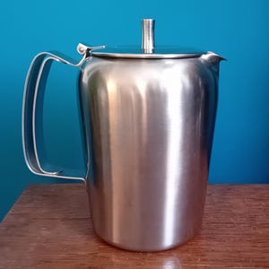 May include: A silver stainless steel teapot with a hinged lid and a handle. The teapot is sitting on a wooden surface.