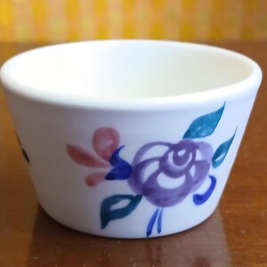 May include: A small, off-white ceramic bowl featuring a hand-painted floral design. The motif consists of a central purple rose with blue and teal leaves, accented by soft pink petals, wrapping around the exterior of the rounded vessel.