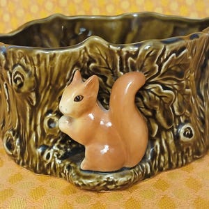 May include: A ceramic planter shaped like a tree stump, with a brown and green glaze. A small, orange squirrel figurine is attached to the front. The planter has a textured surface and is designed to hold small plants or flowers.