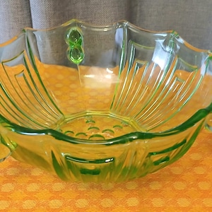 May include: A vintage green glass bowl with a scalloped rim and a fan-like design. The bowl has two rounded handles and decorative accents. The bowl is sitting on an orange surface.