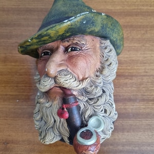 May include: A detailed ceramic bust of an elderly man with a long, curly white beard and a green hat. He is smoking a pipe with a red tassel. The bust is painted with realistic skin tones and textures.