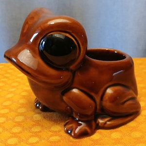 May include: A brown ceramic frog-shaped planter with large, black eyes. The planter has a glossy finish and a small opening for plants or other decorative items. The frog is sitting with its legs tucked beneath its body.