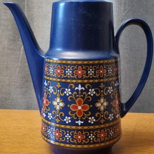 May include: A vintage blue ceramic coffee pot with a decorative floral pattern in red, white, and gold. The pot has a long spout, a curved handle, and a lid. The coffee pot is sitting on a wooden surface.