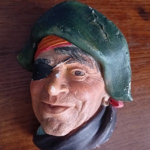 May include: A weathered pirate bust with a green hat, red and orange bandana, and a black eye patch. The pirate has a black scarf and a smiling expression. The bust is painted with realistic details and is set against a wooden background.