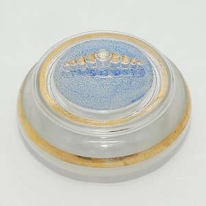 May include: A small, round, white glass jar with a gold rim and a blue and gold lid. The lid has a decorative design of a row of small, gold circles with a larger, white circle in the center.