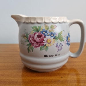 May include: A white ceramic pitcher with a floral design and a scalloped rim. The pitcher has a handle and the words "Mevagissey" printed on the front.