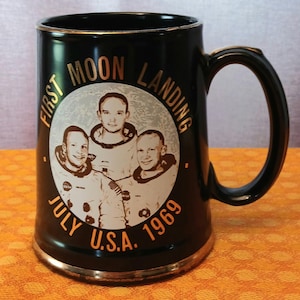 Peut inclure: Mug noir avec bordure dorée, portant l'inscription "FIRST MOON LANDING JULY U.S.A. 1969" autour d'un cercle argenté avec des images de trois astronautes. La tasse a une anse noire et un bord doré.
