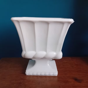 May include: White ceramic planter with a square base and a fluted design. The planter has a pedestal base.