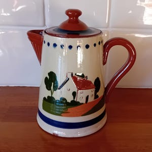 May include: A white ceramic teapot with a red lid and handle. The teapot features a blue and white design with a painted scene of a house and trees.