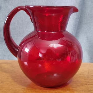 May include: A vibrant red glass jug with a rounded base and a curved handle. The jug features a pouring spout. The glass reflects light, creating highlights and shadows. It is likely used for serving drinks, with a capacity of approximately 1.5 litres.