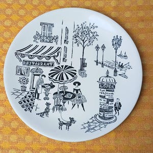 May include: White ceramic plate featuring a black and white illustration of a Parisian street scene. The design includes a restaurant, cafe tables, figures, and a street post with text. The plate has a simple, elegant design.