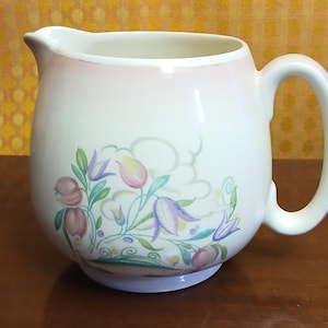 May include: A cream-colored ceramic pitcher featuring a soft pink gradient near the rim and a floral motif on the front. The design includes purple and pink bell-shaped flowers, green leaves, and swirling vines. It has a curved handle and a spout.
