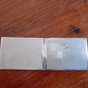 May include: A silver compact with a textured surface and the word "St. Andrews" embossed on the front.