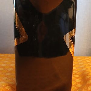 May include: A rectangular black bottle with gold accents. The bottle has a glossy finish and a label that reads "DE LUXE". The bottle is sitting on a yellow patterned surface.