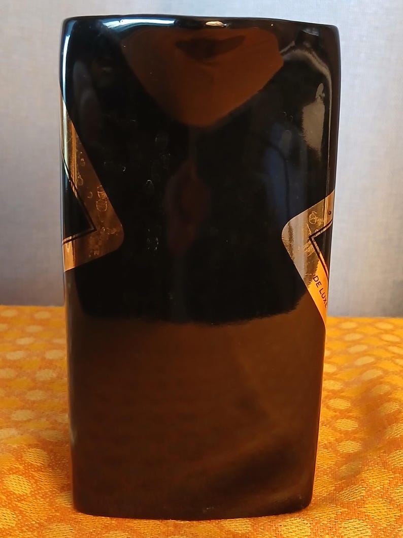 May include: A rectangular black bottle with gold accents. The bottle has a glossy finish and a label that reads "DE LUXE". The bottle is sitting on a yellow patterned surface.