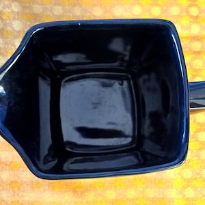 May include: A dark blue ceramic mug with a square body and a handle. The mug has a glossy finish and a unique, angular design. The mug is empty and sits on a yellow and white patterned surface.