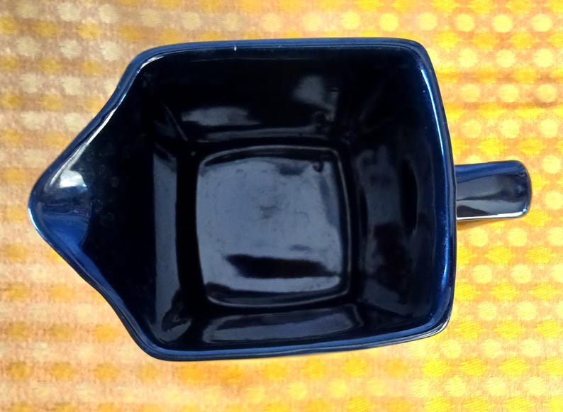 May include: A dark blue ceramic mug with a square body and a handle. The mug has a glossy finish and a unique, angular design. The mug is empty and sits on a yellow and white patterned surface.