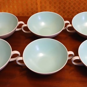 May include: A set of six matching soup bowls arranged in two rows of three. Each bowl features a smooth, pale mint green interior and a contrasting white exterior, complete with two small, curved handles on opposite sides for easy gripping.