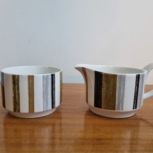 May include: A white sugar bowl and creamer set with a black, gold and grey striped pattern.