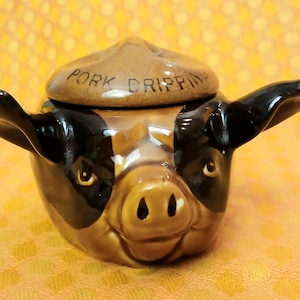 May include: A ceramic pig-shaped jar for pork dripping, with a lid. The jar is brown, black, and tan, with the words "PORK DRIPPING" on the lid. The pig has large ears and a smiling expression. A kitchenware item.