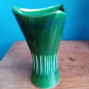 May include: A green ceramic vase with a unique, asymmetrical design. The vase has a wide, flared opening and a narrow base with vertical lines.