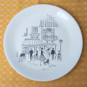 May include: Round white ceramic plate featuring a black and white illustration of a European street scene. The design includes figures, buildings, and awnings, giving a vintage feel. The plate has a glossy finish.