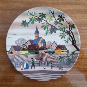 May include: A round ceramic plate featuring a colourful painted scene of a village with houses, a church, and a tree. The plate has a brown background with a white rim.