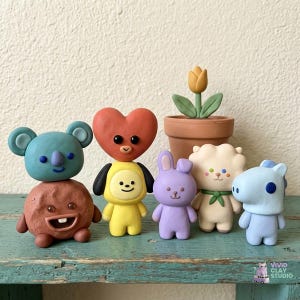 May include: A collection of colorful clay figurines, including a koala, a heart-shaped character, a yellow figure, a purple bunny, a white sheep, and a blue horse. A small terracotta pot with a yellow tulip is also present. The figurines are displayed on a teal surface.
