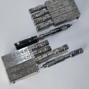 Op de afbeelding: Een assortiment metalen pennen en pennendoosjes. De pennen hebben een strak, industrieel ontwerp met ingewikkelde details en gekleurde accenten. De doosjes zijn rechthoekig met gegraveerde bloempatronen. De pennen en doosjes zijn op een wit oppervlak geplaatst.
