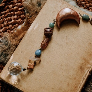 May include: A handcrafted celestial-themed pendant necklace with a crystal ball, wooden beads, and a crescent moon charm. The necklace is displayed on a vintage book with a worn cover, set against a textured background.