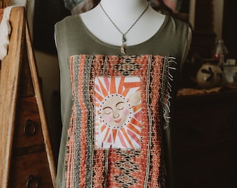 May include: Olive green tank top with a colorful, stitched patch featuring a smiling sun design. The patch is framed by patterned fabric in shades of red, orange, and gray. A delicate necklace with a pendant is also visible.