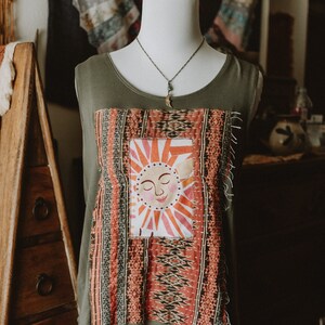 Puede incluir: Camiseta sin mangas verde oliva con un parche cosido colorido con un diseño de sol sonriente. El parche está enmarcado por tela estampada en tonos rojos, naranjas y grises. También se ve un delicado collar con un colgante.