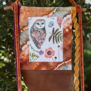 May include: A decorative wall hanging featuring an owl illustration, flowers, and foliage on a white background. The banner has a brown suede bottom, orange and purple accents, and a macrame cord. The banner is approximately 18 inches long.