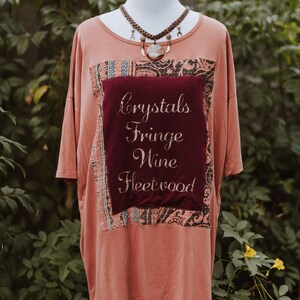 May include: A coral-colored t-shirt with a burgundy square patch featuring the words "Crystals Fringe Wine Fleetwood" in cursive. The patch is framed by patterned fabric. A necklace with a pendant is displayed.
