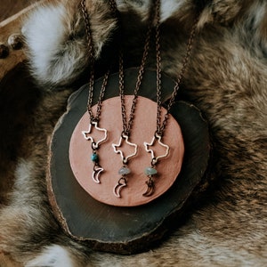 May include: Three copper necklaces with a Texas state outline pendant and a crescent moon charm. Each necklace features a different colored gemstone: turquoise, amethyst, and green aventurine.