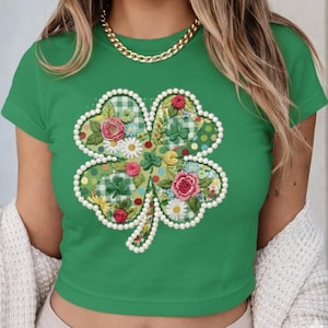 May include: A green crop top with a four-leaf clover design. The clover is decorated with floral patterns, including roses and daisies, and edged with white pearls. The top has short sleeves and a round neckline.