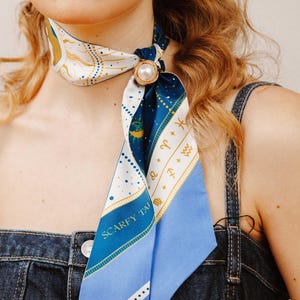 May include: A silk scarf with a celestial design, featuring zodiac symbols and the text "SCARFY TALES". The scarf is primarily blue and white, with gold accents and a pearl-adorned ring clasp.