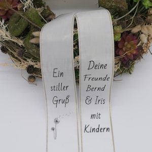 Könnte beinhalten: Ein weißes Band mit goldenem Rand und schwarzer Fransen, mit dem Text "Ein stiller Gruß" und "Deine Freunde Bernd & Iris mit Kindern." Ein grünes Banner darüber lautet "Trauerschleifenmanufaktur.de Aus Tradition seit 1947."